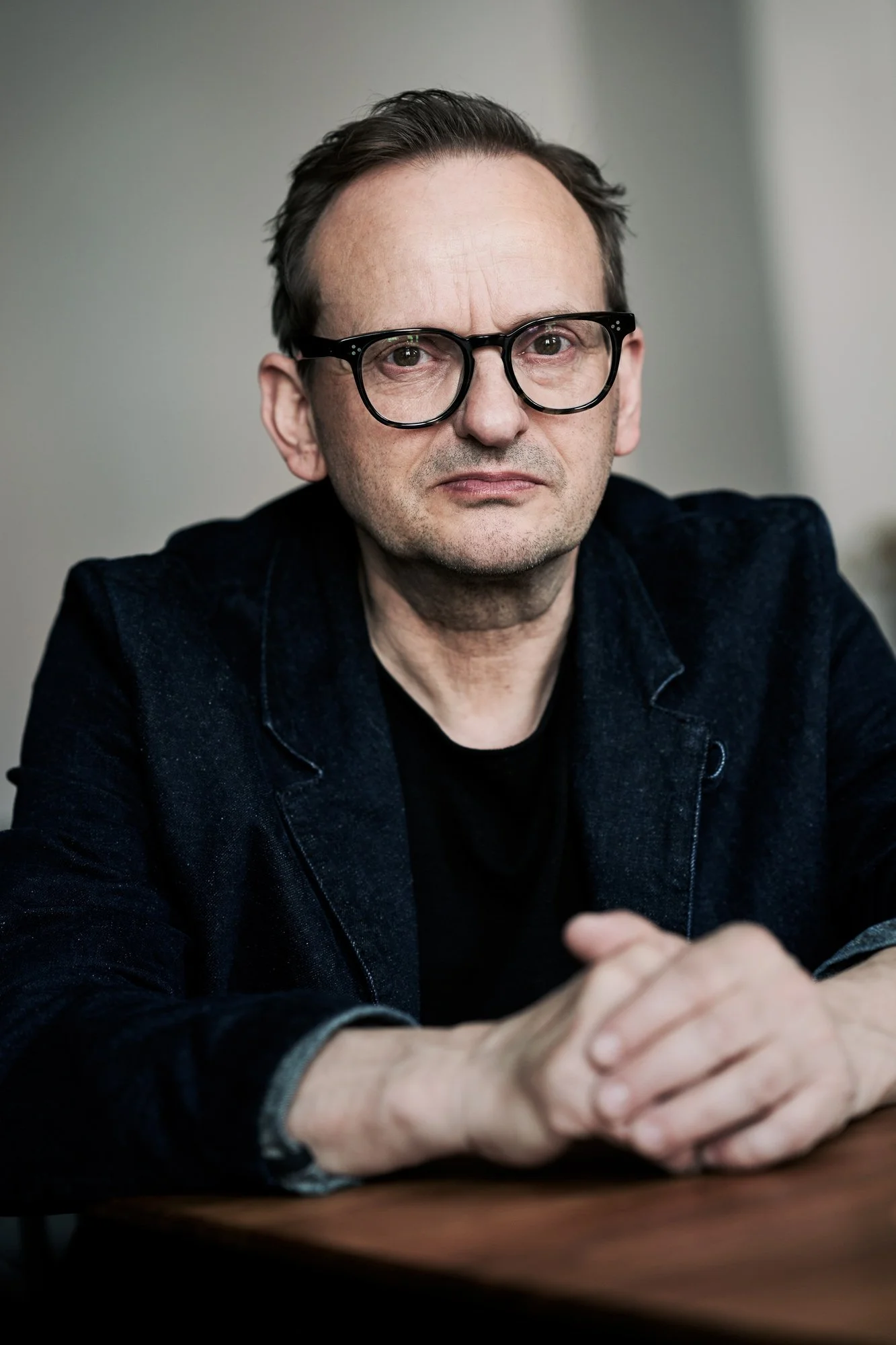 Porträtaufnahme des Schauspielers Milan Peschel mit Brille im dunkelblauen Jeansblazer, an einem Holztisch sitzend mit gefalteten Händen, direkter, ernster Blick in die Kamera, fotografiert von Koszki Photography Berlin.
