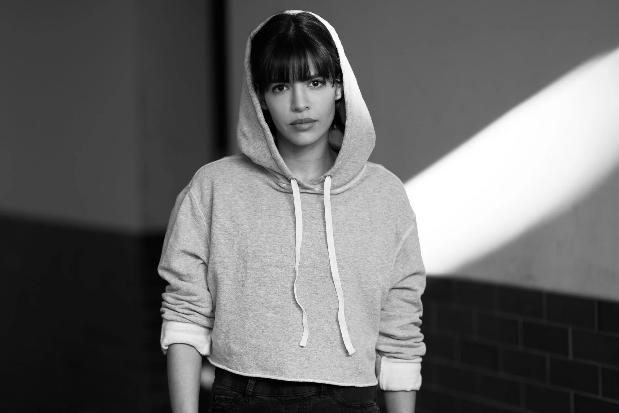 Schwarz-Weiß-Porträt der Schauspielerin Jasmin Lord im Kapuzenpullover mit aufgesetzter Kapuze, mit ernstem Blick in die Kamera vor einer Wand mit einem diagonalen Lichtstreifen, fotografiert von Koszki Photography Berlin.