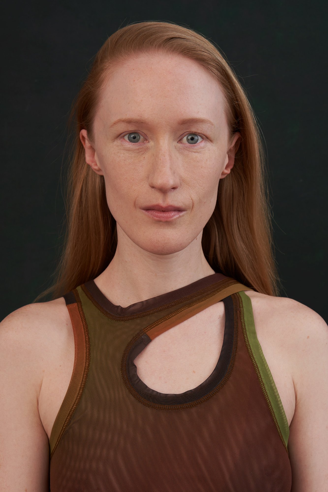 Studioportrait der Künstlerin und Schauspielerin Britta Thie mit langen roten Haaren im braun-grünen Mesh-Top mit Cut-out am Ausschnitt, frontal mit ruhigem, direktem Blick vor dunklem Hintergrund, fotografiert von Koszki Photography Berlin.