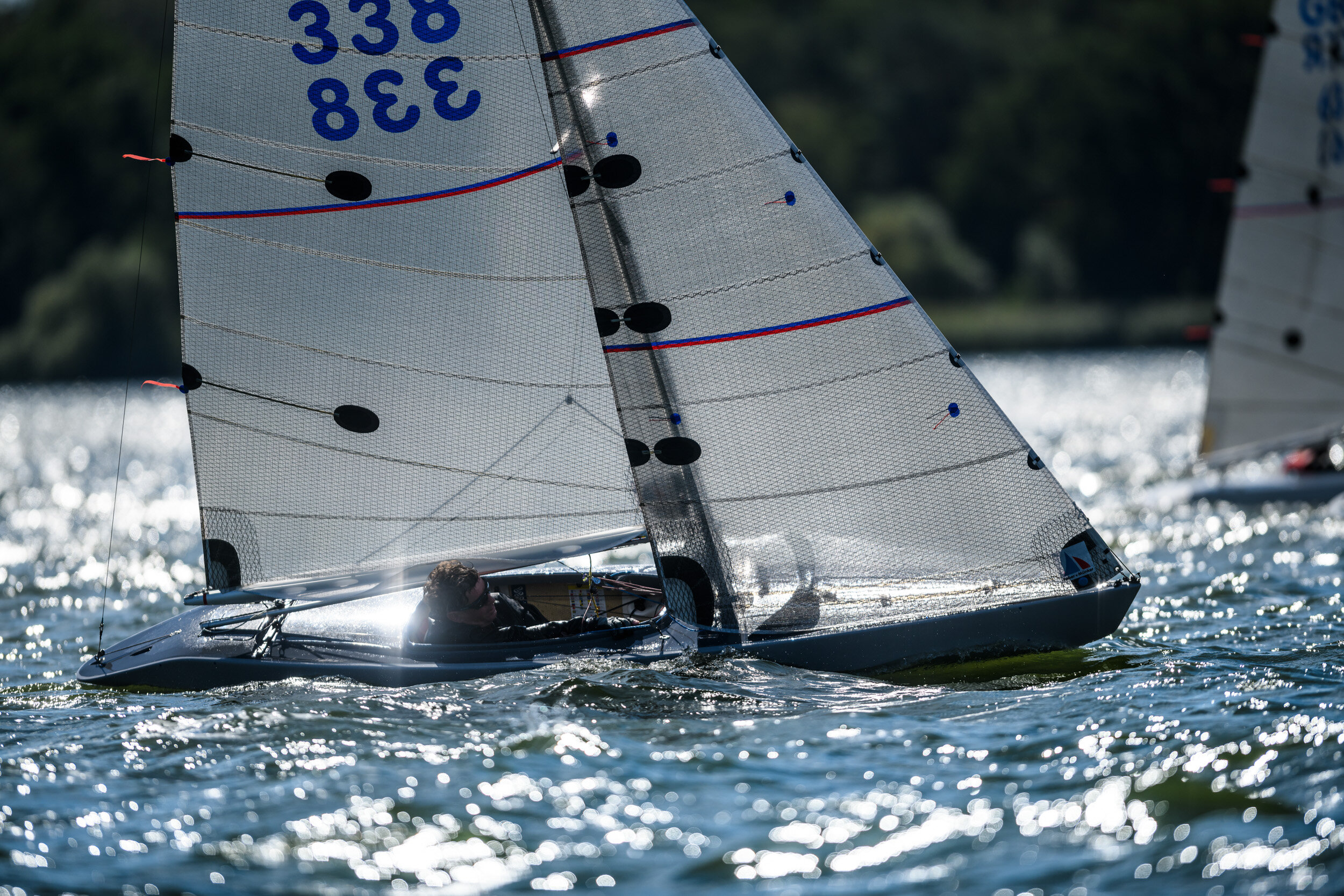 A 2.4mR sailboat racing on a body of water with another boat visible in the background, the boat has a high-tech sail with numbers, and a person wearing sunglasses is sitting inside.