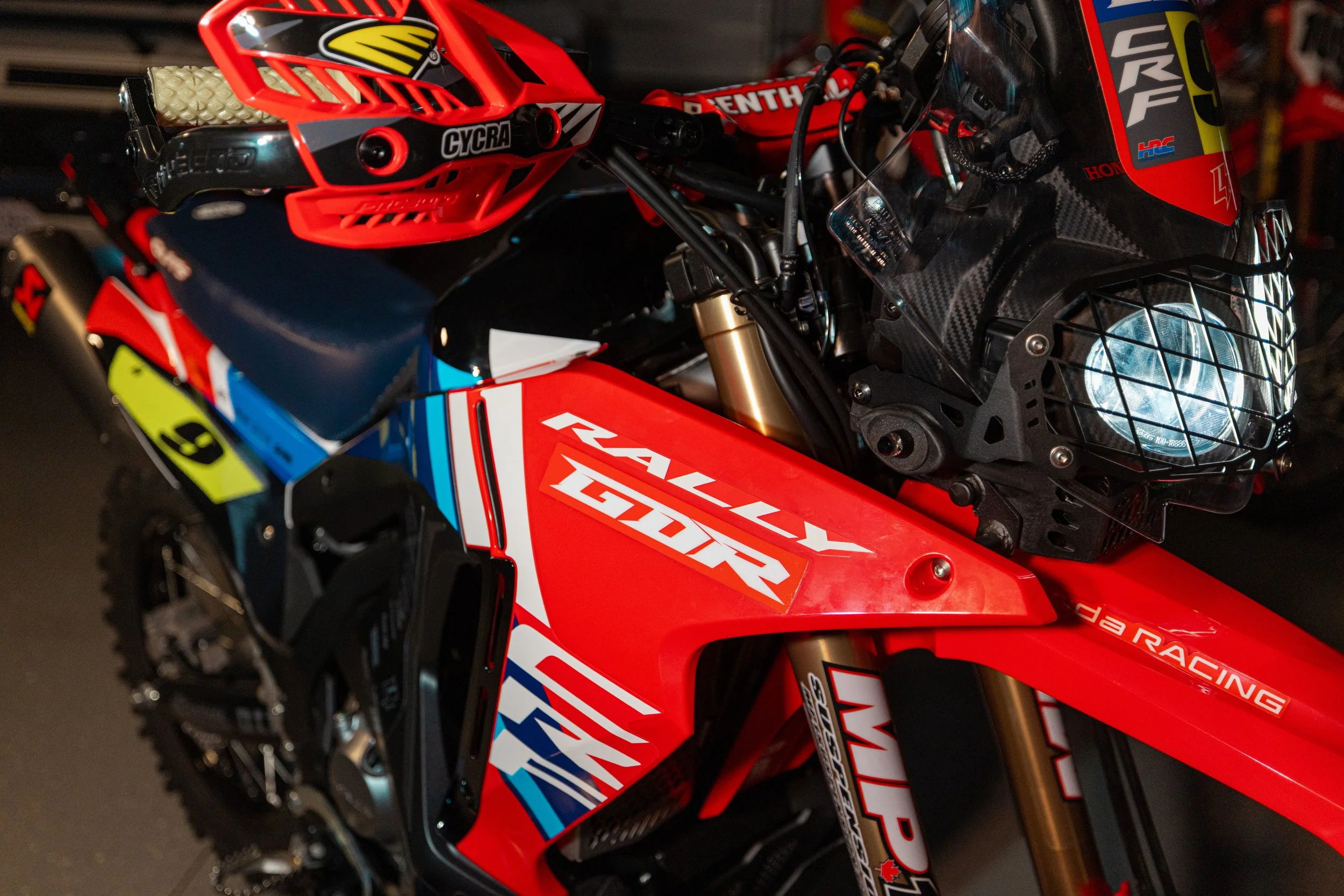 Close-up of a red, black, and blue racing motorcycle with a helmet resting on the seat. The motorcycle features various brand labels, including 'Rally RTR,' 'Honda,' and 'WP.' The front headlight is protected by a metal cage.