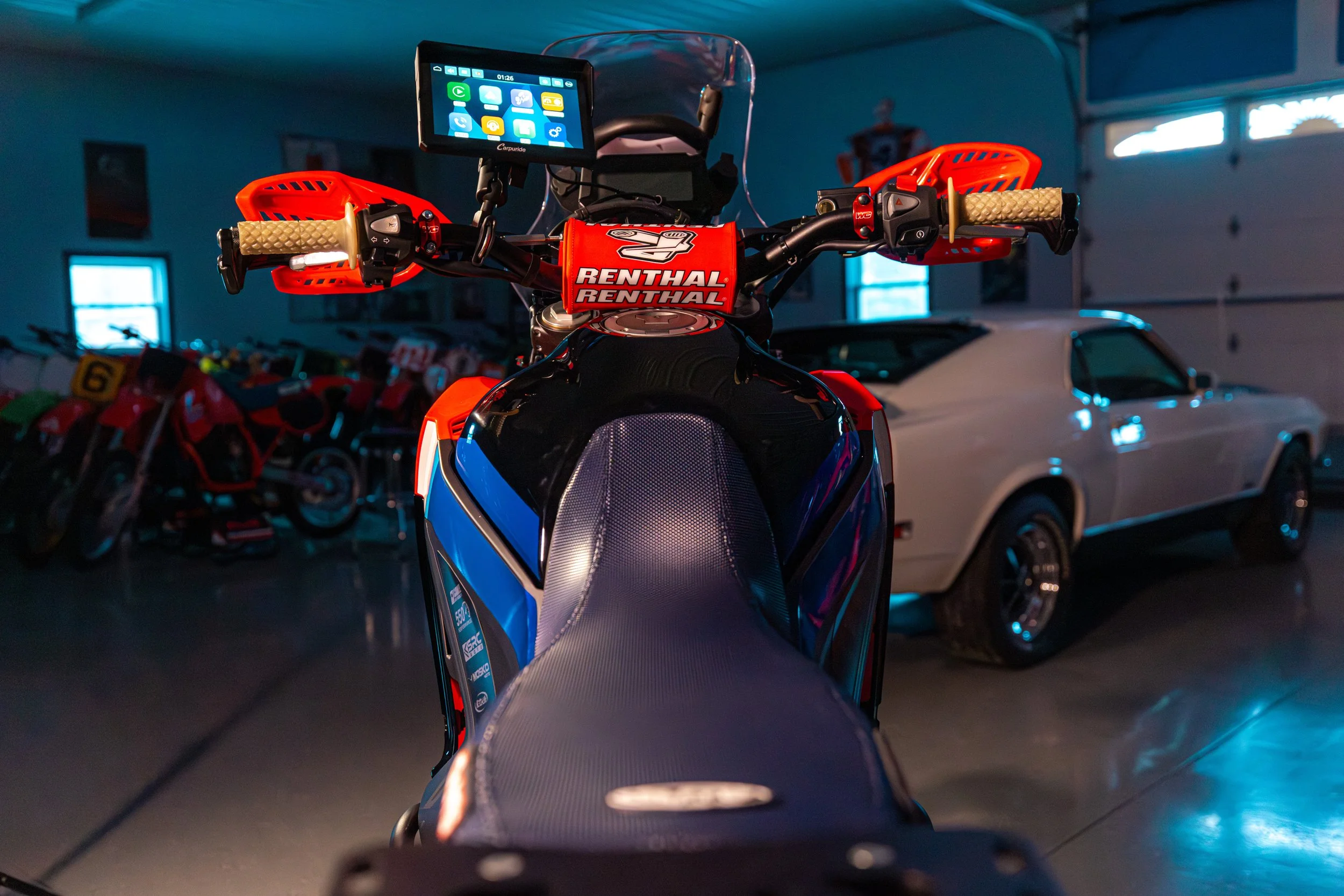 A dirt bike with a blue and black seat, orange handlebars, and a transparent windscreen, parked indoors with a vintage white car and several other bikes in the background.