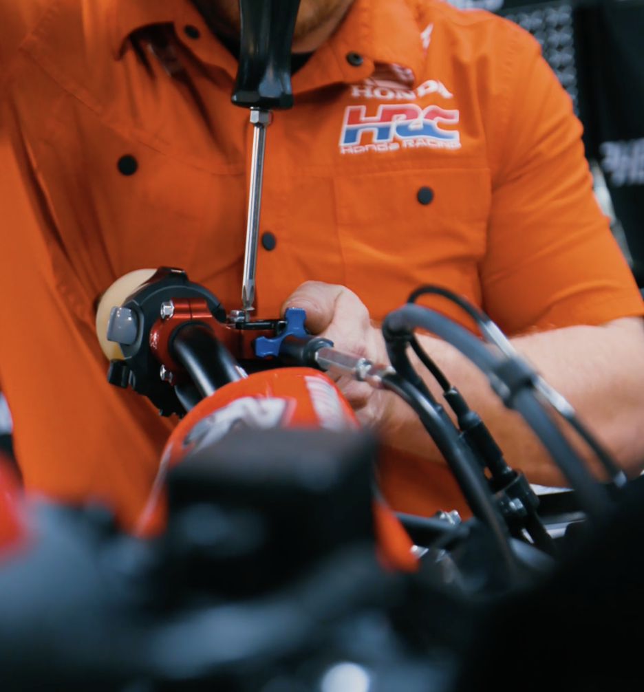 Person in an orange Honda HRC racing shirt working on a motorcycle handlebar in a garage or workshop.