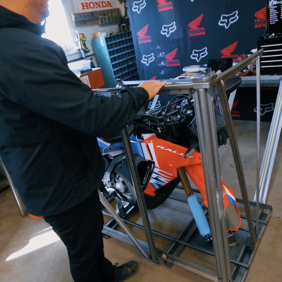A person standing next to a partially assembled orange and black Honda motorcycle, inside a workshop with Honda and Fox logos on the backdrop.