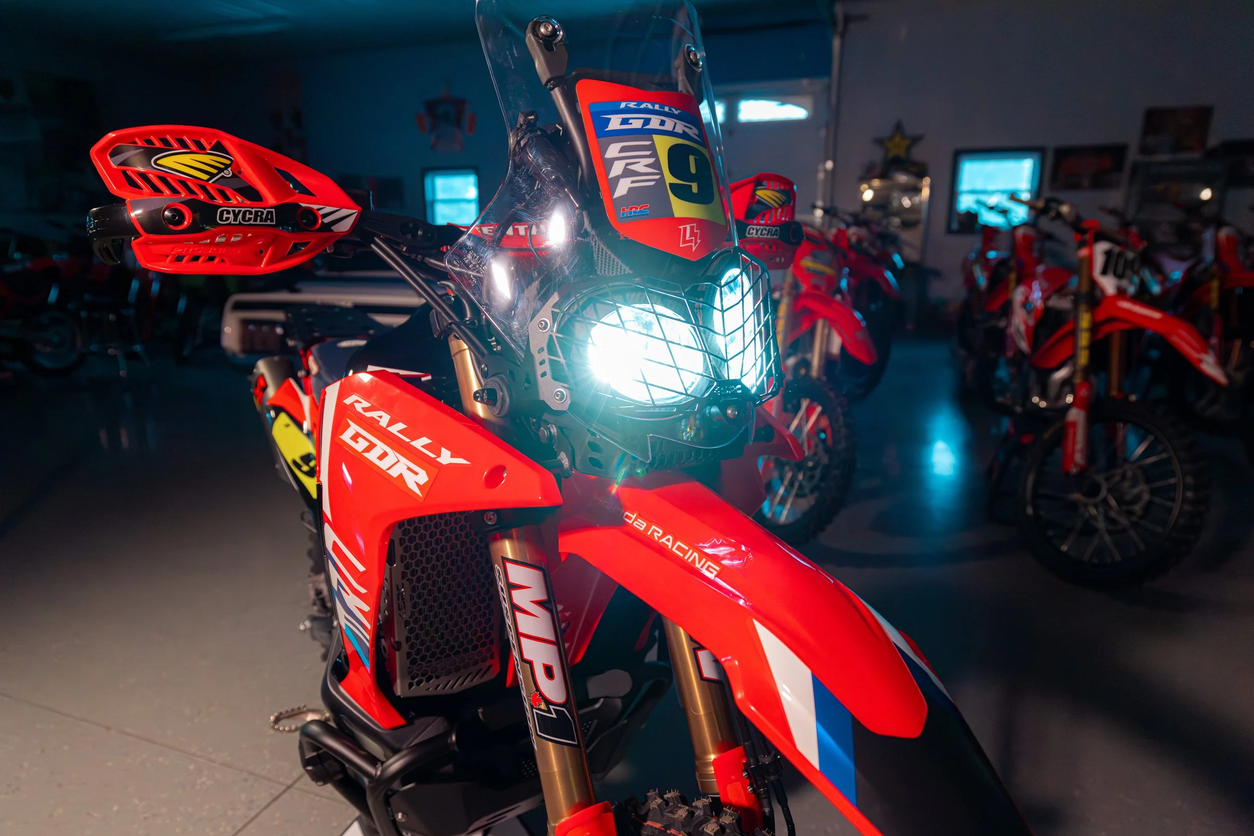 Red motocross bike in a showroom with other bikes in the background, featuring a front headlight, racing decals, and a helmet resting on the handlebar.