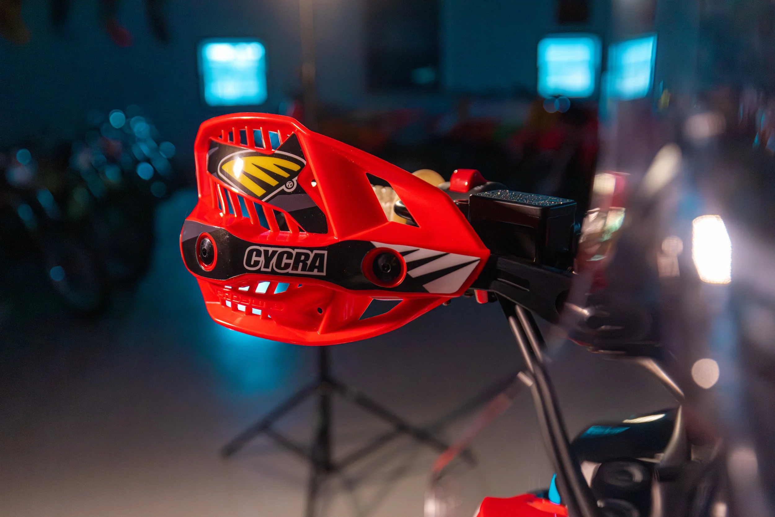 Close-up of a red motocross helmet with black and white graphics, mounted on a stand in a dimly lit room with blurred background.