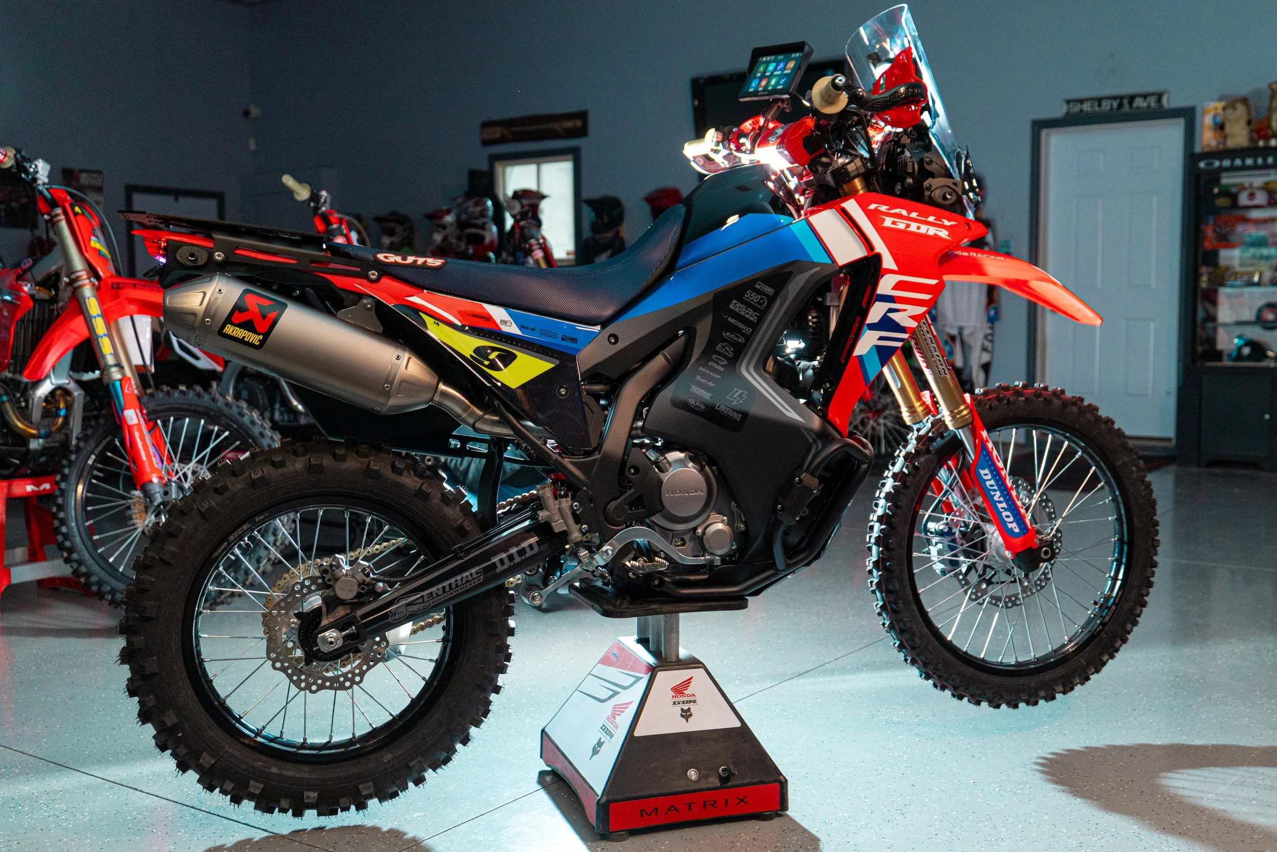 A Honda extreme off-road motorcycle on display indoors, with red, blue, and black graphics, knobby tires, and a tall suspension, surrounded by similar bikes in the background.