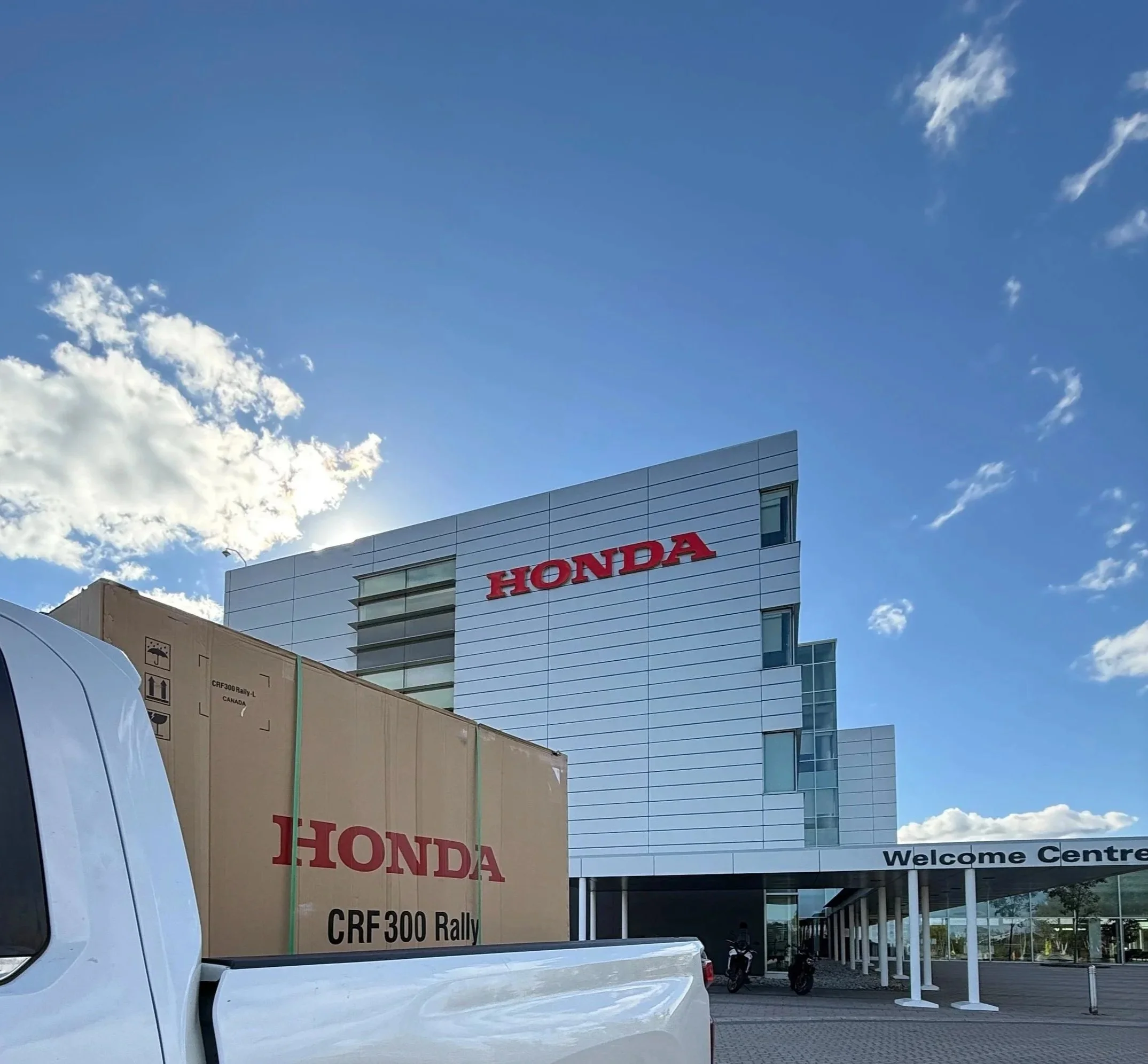 Exterior view of a modern Honda building with a large red Honda sign, a welcome center, and a white truck with a Honda CRF300 Rally motorcycle box in the foreground.