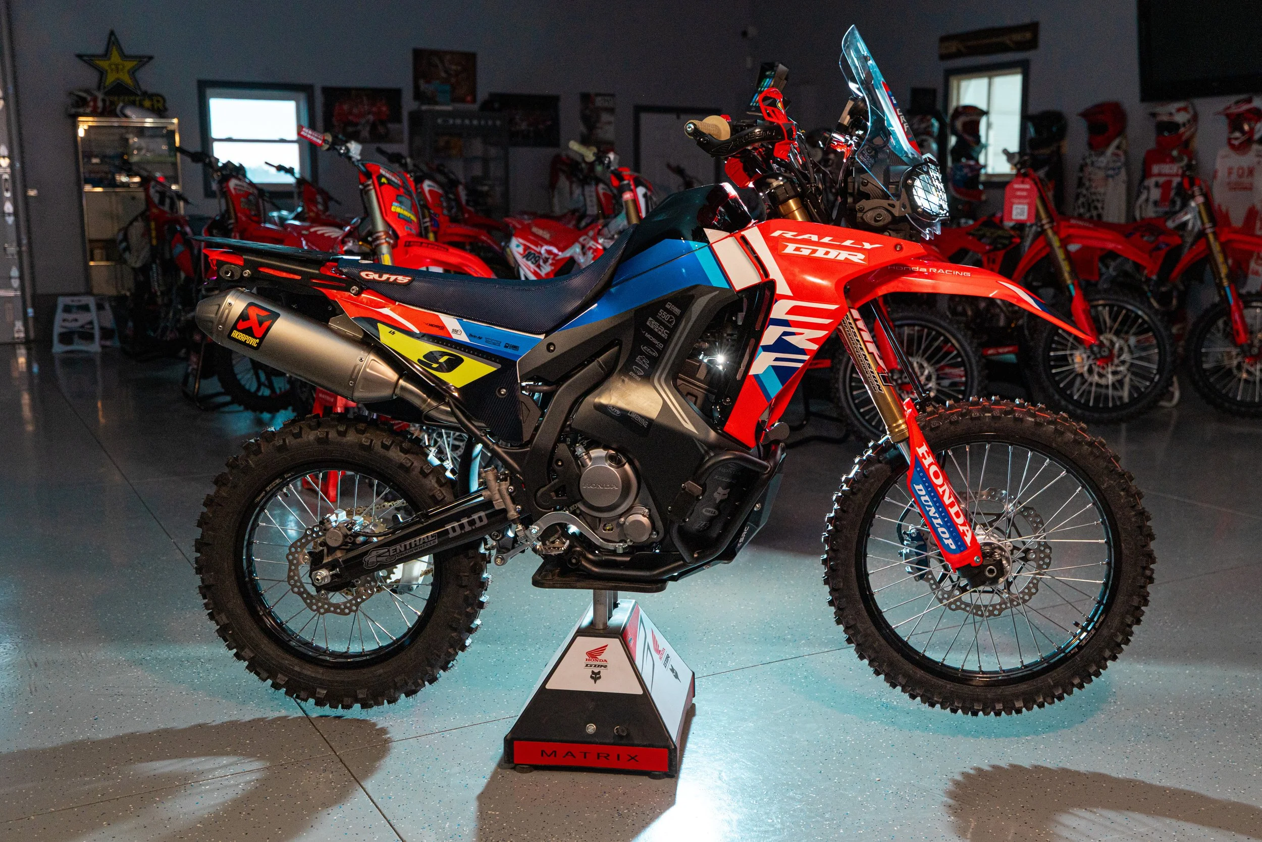 A red, blue, and black Honda off-road motorcycle displayed on a stand inside a showroom, with other motorcycles and motocross helmets in the background.