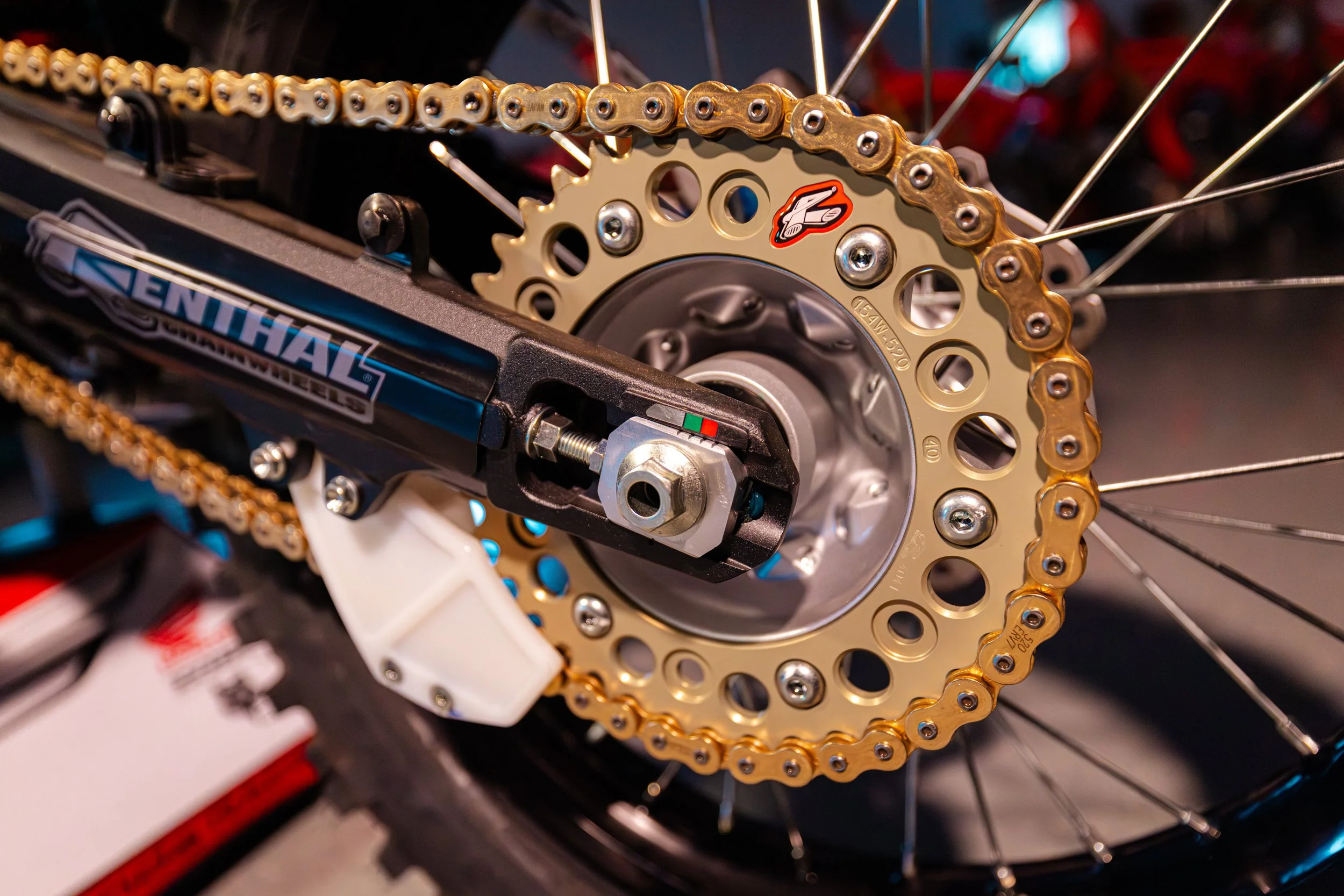 Close-up of a motorcycle rear wheel, featuring a gold sprocket, chain, and black suspension component with a logo.