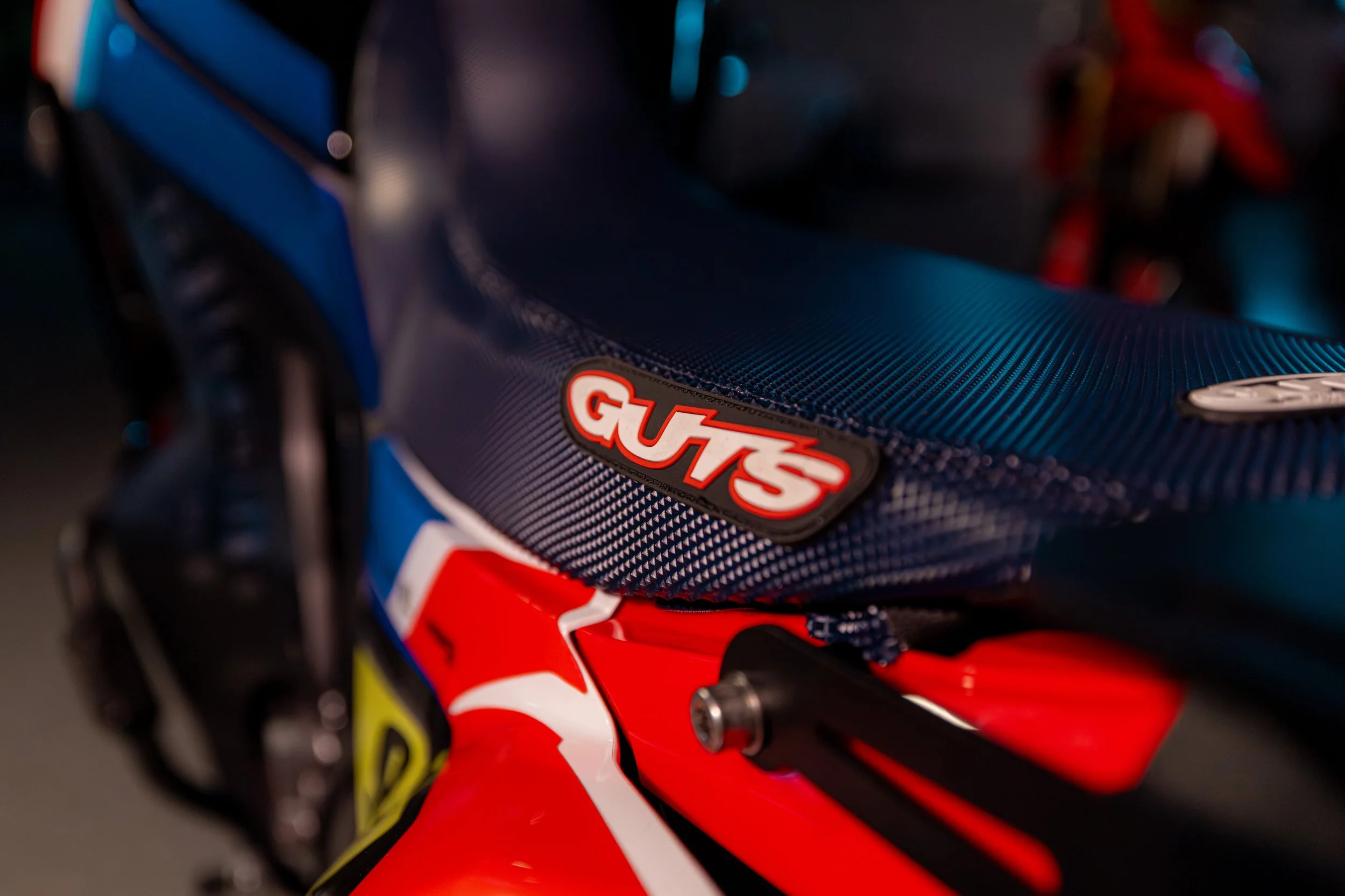 Close-up of a motorcycle seat with a Guts sticker, featuring a textured blue seat and red, white, and blue body parts.