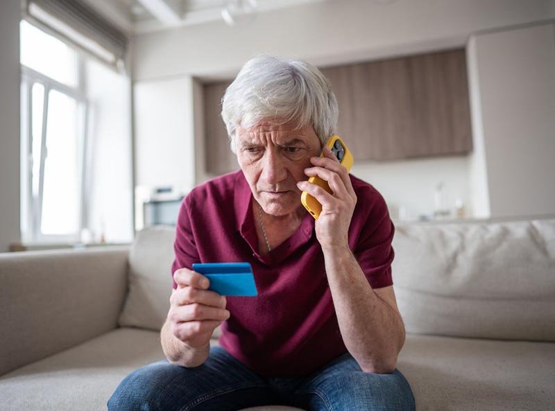 Person sitting on a couch holding a blue credit card and a yellow smartphone, appearing to make a phone call in a modern living room.