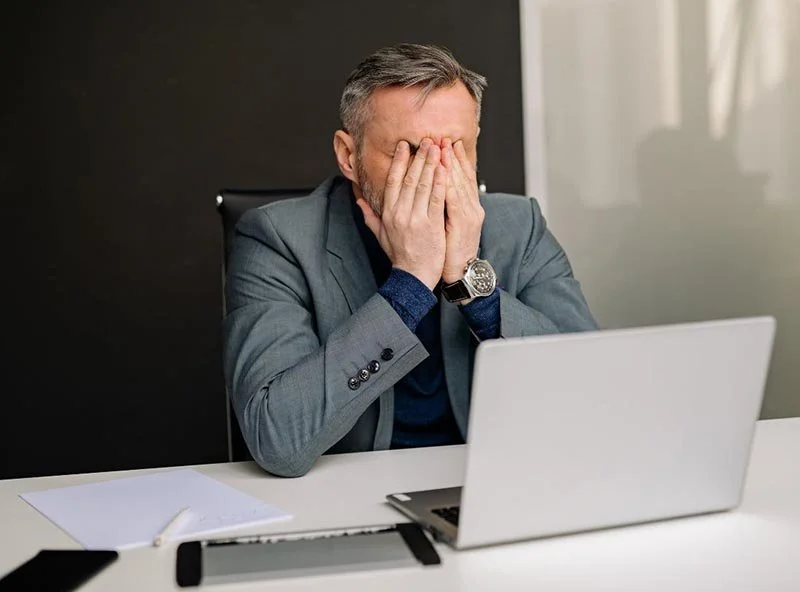 Person seated at a desk with an open laptop, hands covering their face, papers and a keyboard on the desk.