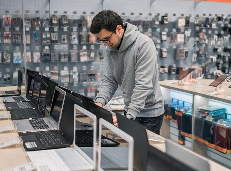 Person examining laptops on display in an electronics store with shelves of accessories in the background.