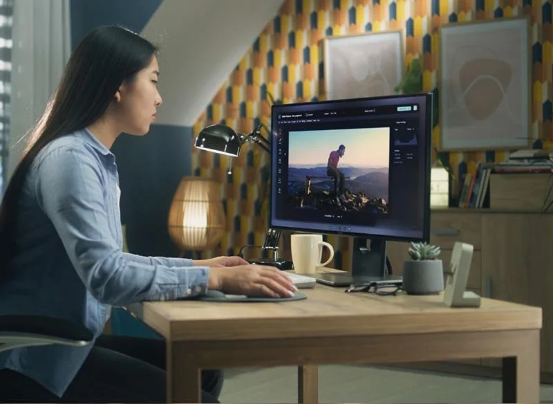Person working at a desk on a computer, editing a photo of someone standing on a mountain at sunset.