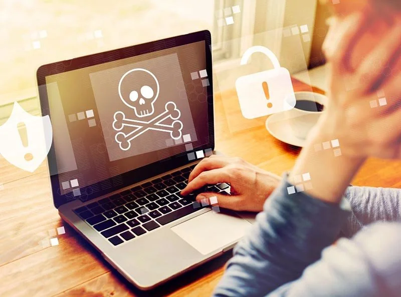 Person using a laptop displaying a skull and crossbones warning symbol, indicating a malware or cybersecurity threat, with security alert icons overlaid.