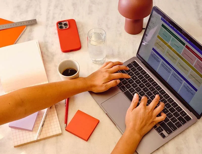 Hands typing on a laptop at a desk with a smartphone, notebook, coffee, and office supplies
