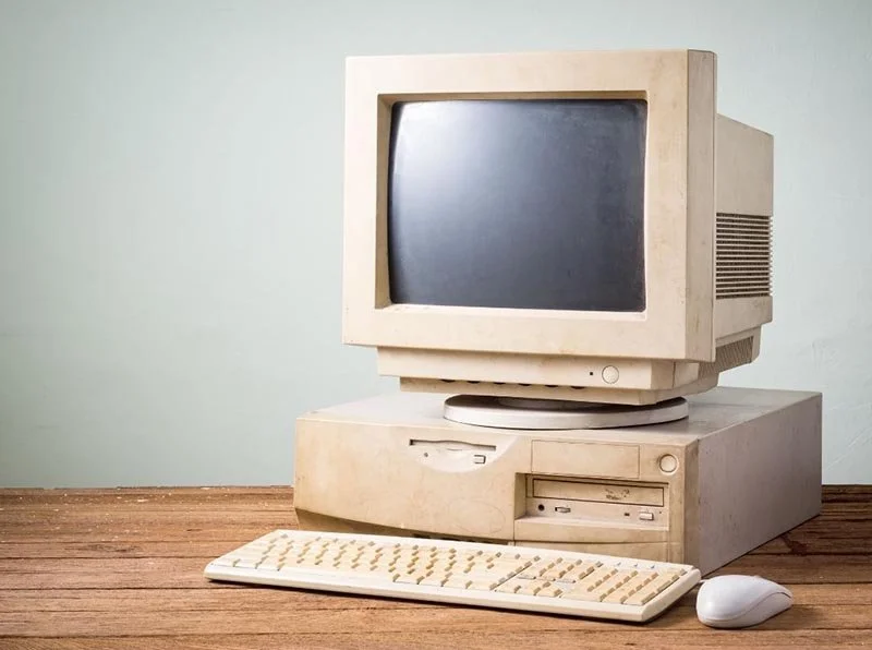 Old beige desktop computer with CRT monitor, floppy disk drive, keyboard, and mouse sitting on a wooden desk.