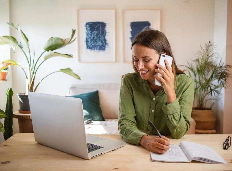 Person working from a home office, talking on a smartphone while taking notes beside an open laptop at a desk.