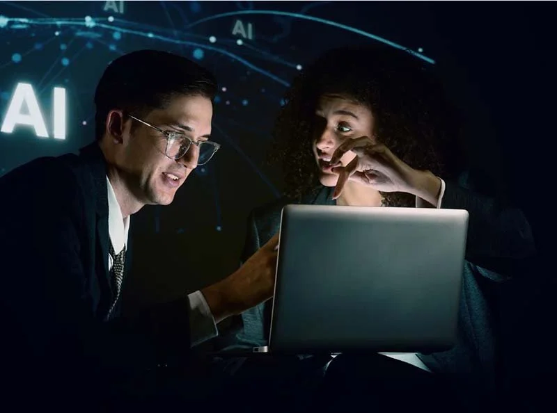 Two professionals working on a laptop with an AI-themed digital display in the background.
