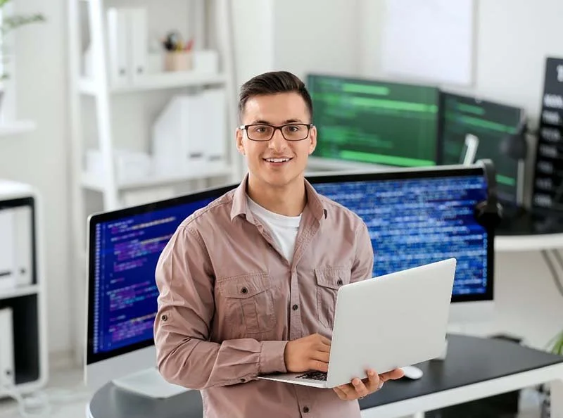 Software professional working on a laptop with multiple monitors displaying code in a modern office workspace.