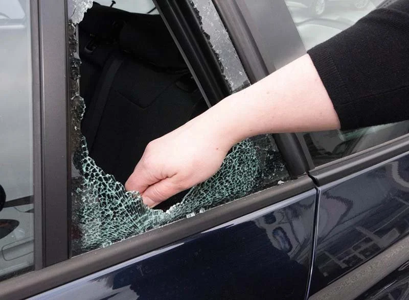 Broken car window with a hand reaching inside the vehicle, representing theft, physical security risks, and the importance of protecting valuable assets.