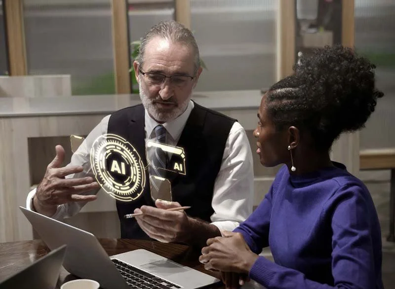 Two professionals seated at a conference table reviewing AI technology concepts on a laptop, with a digital artificial intelligence interface overlay displayed between them.