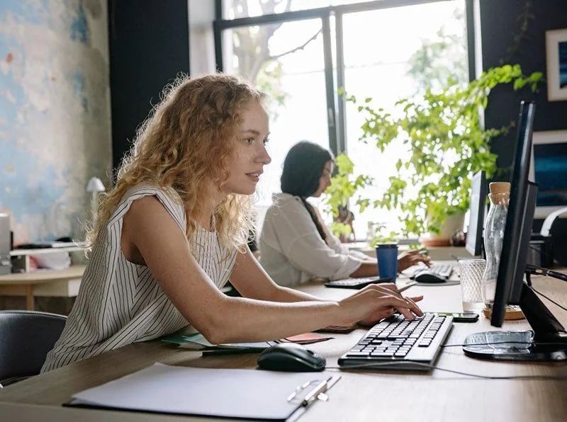 Two people working at desktop computers at a shared desk in a bright office with plants, notebooks, and office supplies.