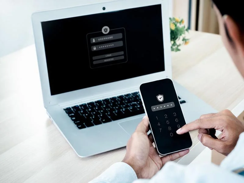 Person using a smartphone to enter a security code while a laptop displays a login screen in the background.