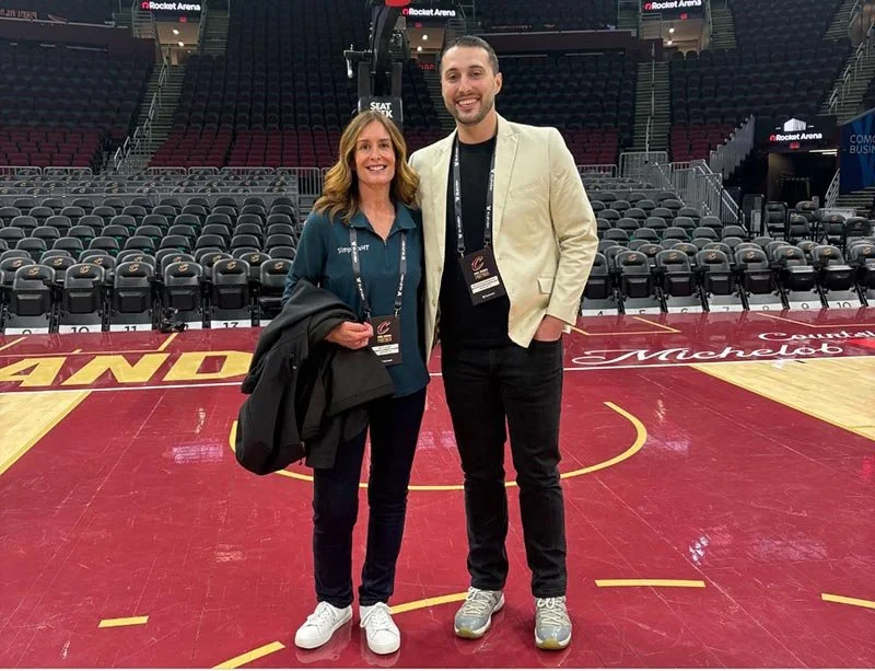 Two people standing on a basketball court inside an empty arena, wearing event badges, with rows of seats visible in the background.