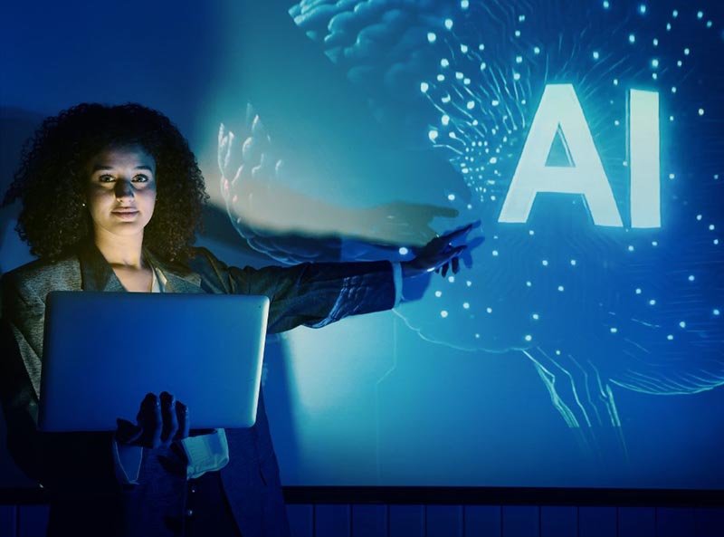 Person holding a laptop and pointing toward a large screen displaying the letters “AI” with a digital brain graphic in the background.
