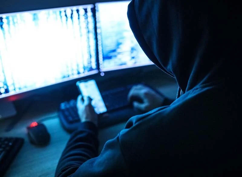 Person in a hooded sweatshirt using a smartphone at a computer workstation with multiple monitors.