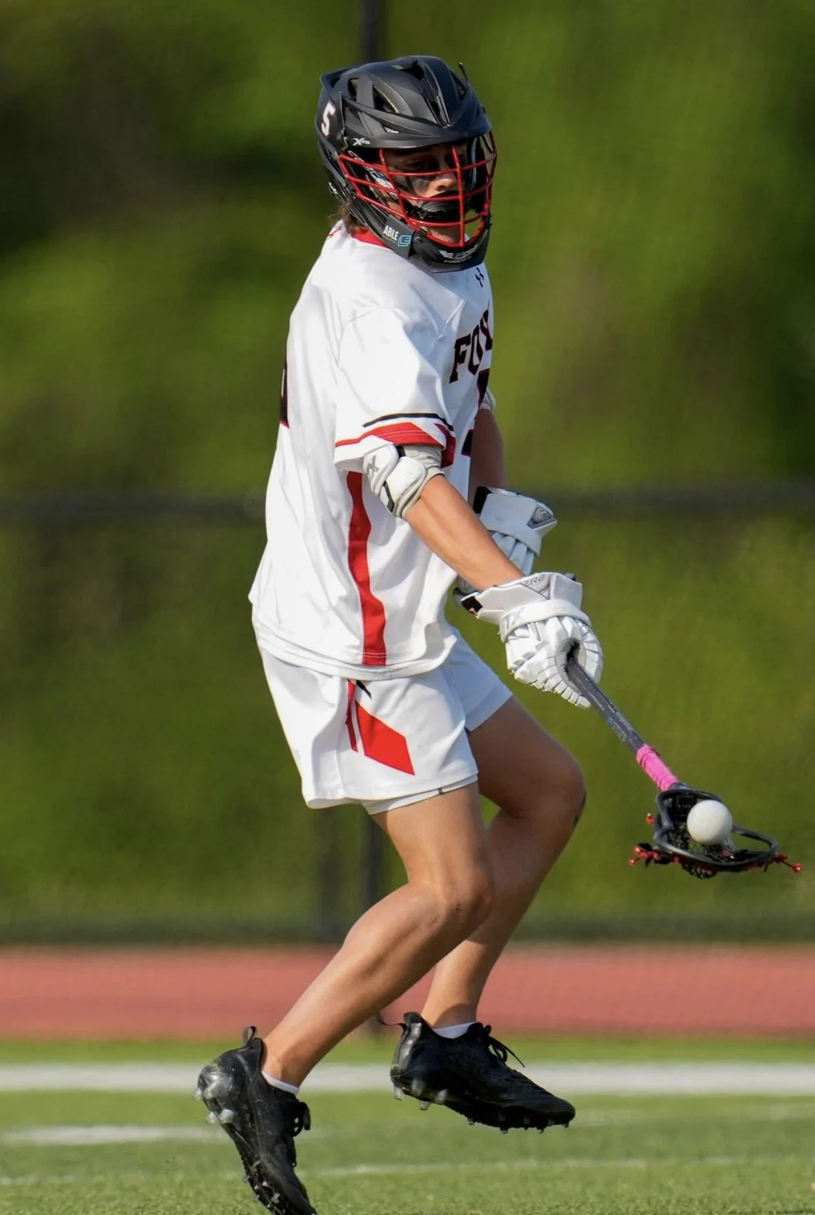 A person playing lacrosse on a field, wearing a black helmet and white sports uniform, holding a lacrosse stick with a ball in the net, mid-air in action.