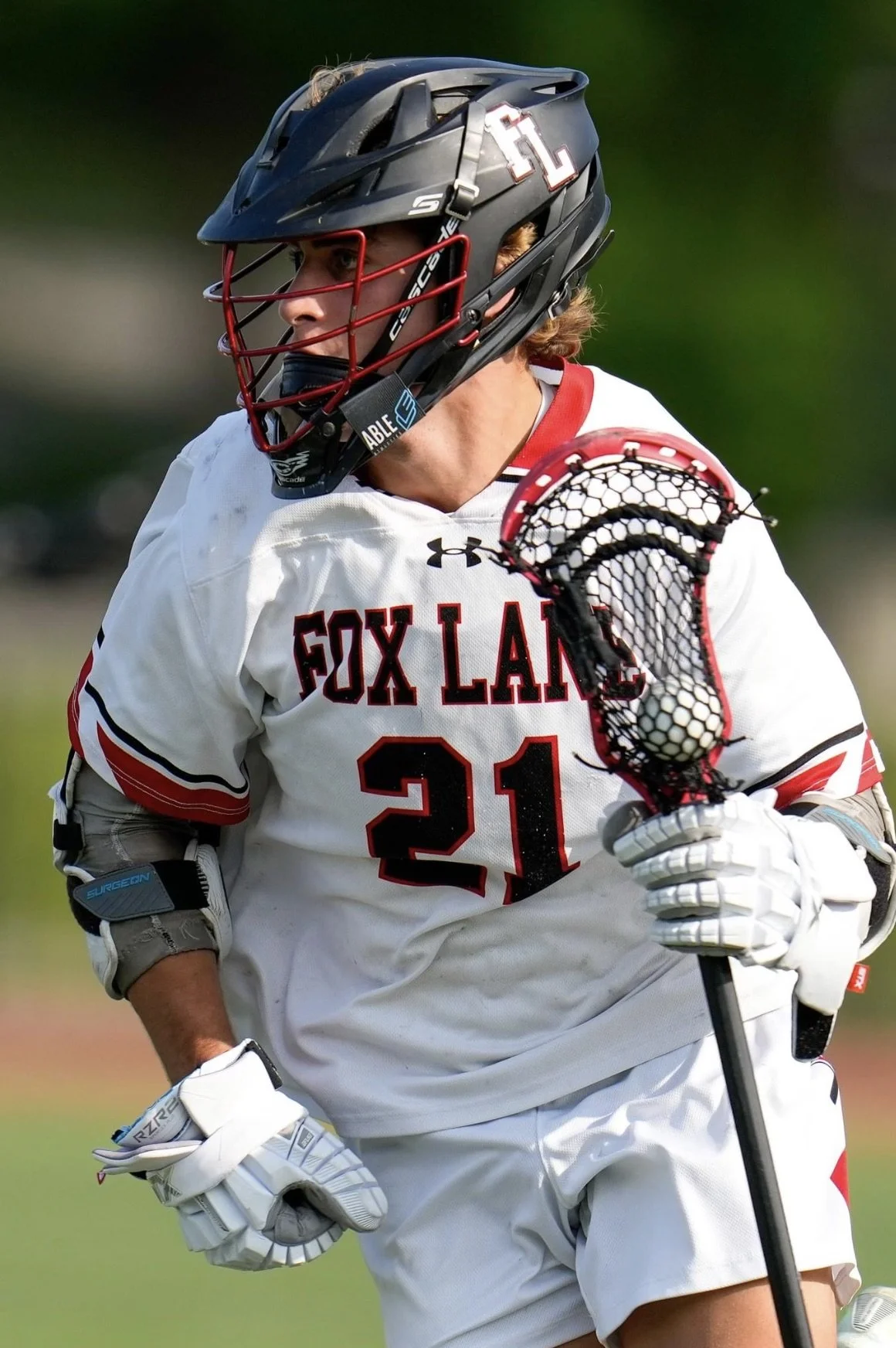 A lacrosse player in a white jersey with red and black accents, wearing a black helmet with a red face mask, holding a lacrosse stick with a mesh net, during a game.