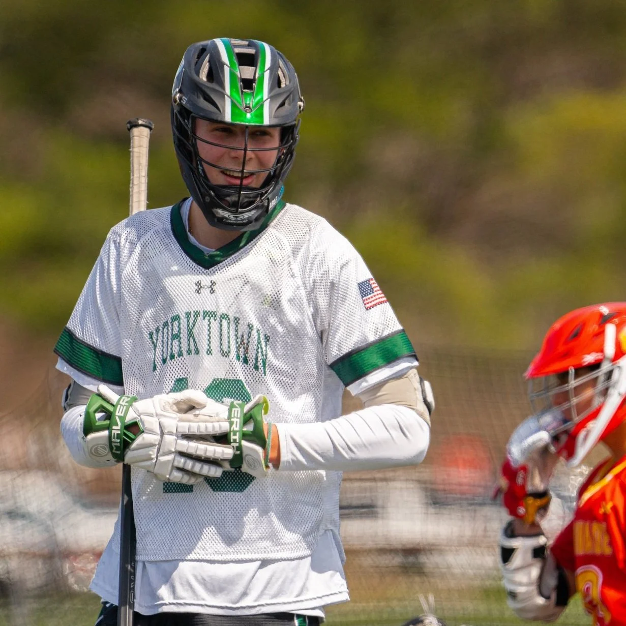 A young male lacrosse player wearing a white Yale University jersey, green helmet with black face guard, and gloves, holding a lacrosse stick, smiling, with a red helmeted player in the background.