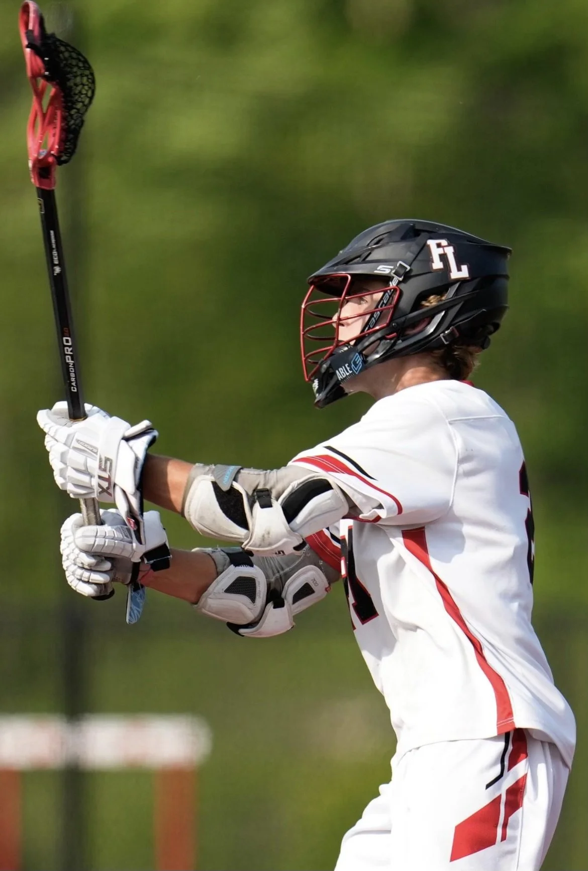 A baseball player in a white and red uniform practicing with a lacrosse stick, wearing a black helmet with a red face guard and white gloves, set outdoors against a green background.
