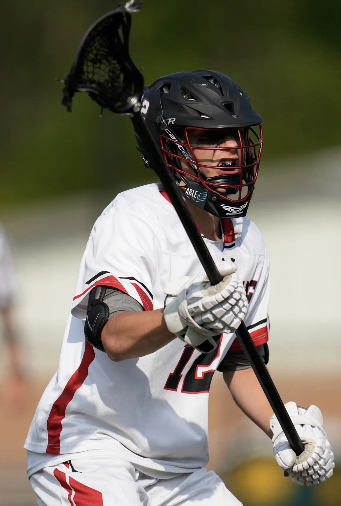 A lacrosse player in a white jersey and black helmet with a red faceguard, holding a lacrosse stick.