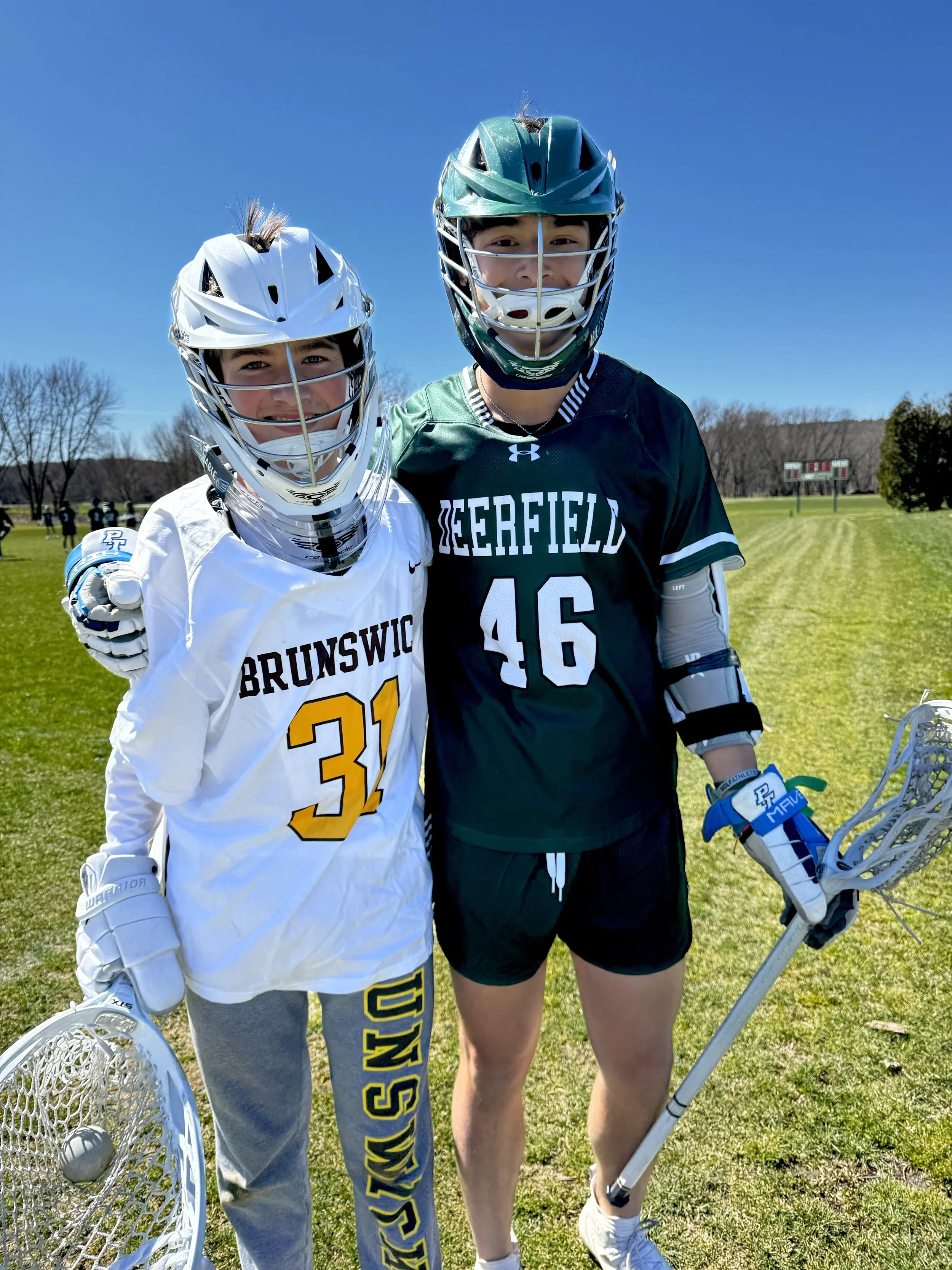 Two male lacrosse players in helmets and uniforms smiling on a grassy field, with one holding a lacrosse stick.