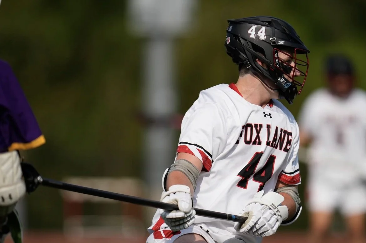 A lacrosse player wearing a black helmet and white jersey with red and black accents, number 44, with 'FOX LANE' written on it, holding a lacrosse stick during a game.