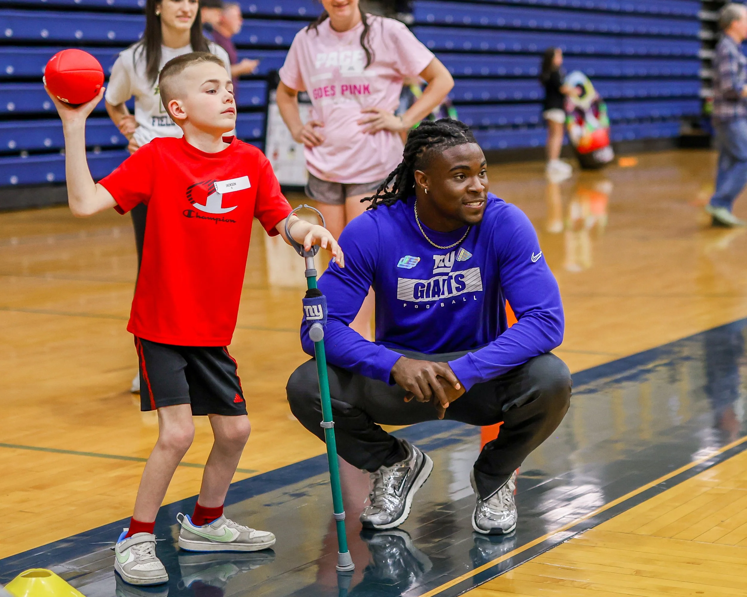 Two children in wheelchairs with lacrosse sticks and a man crouching in the middle holding a white hockey jersey with a lacrosse logo and the words 'Able 6 Athletics' inside a gymnasium.