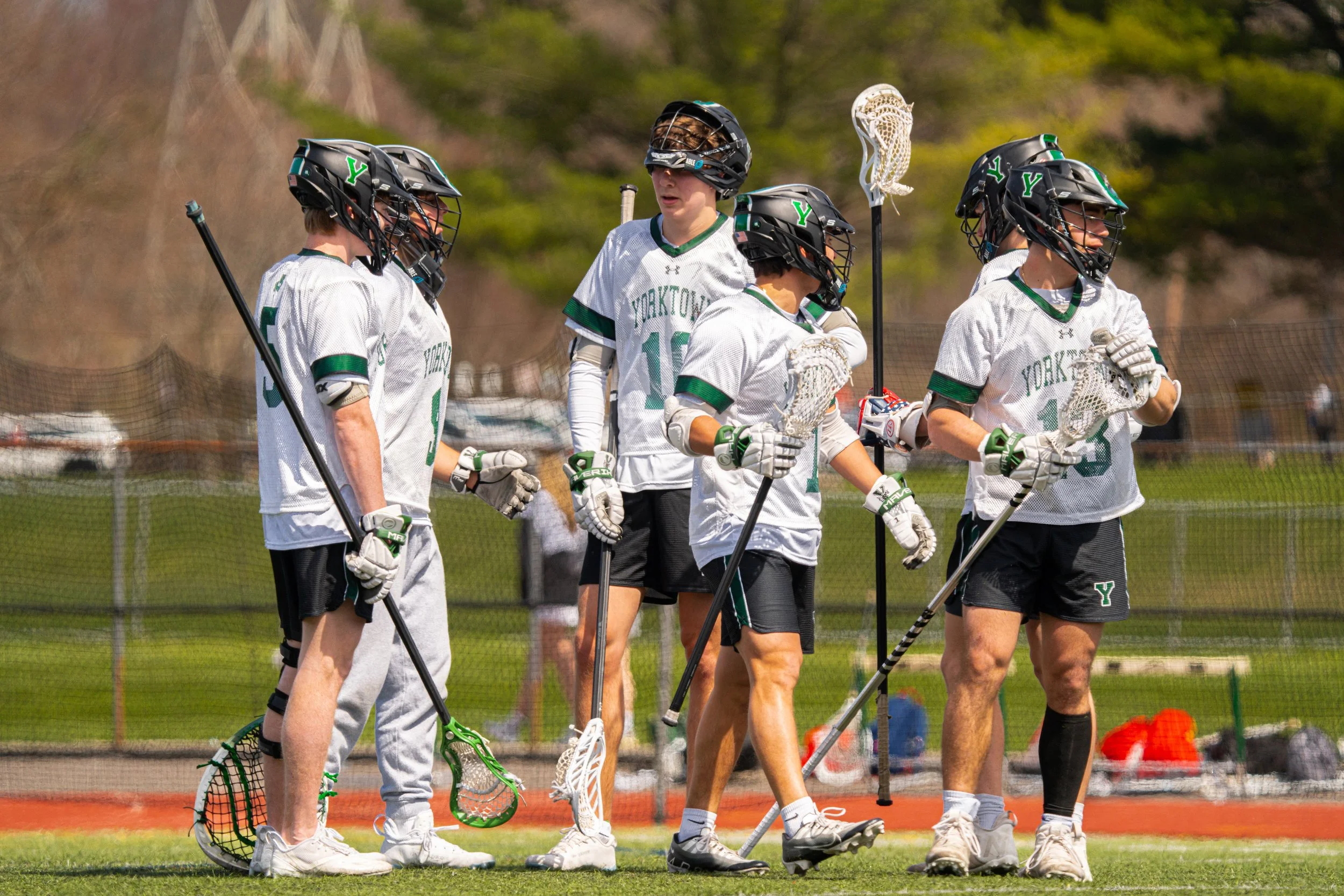 A group of five lacrosse players in white jerseys with green accents and black helmets standing together on a field during a game or practice.