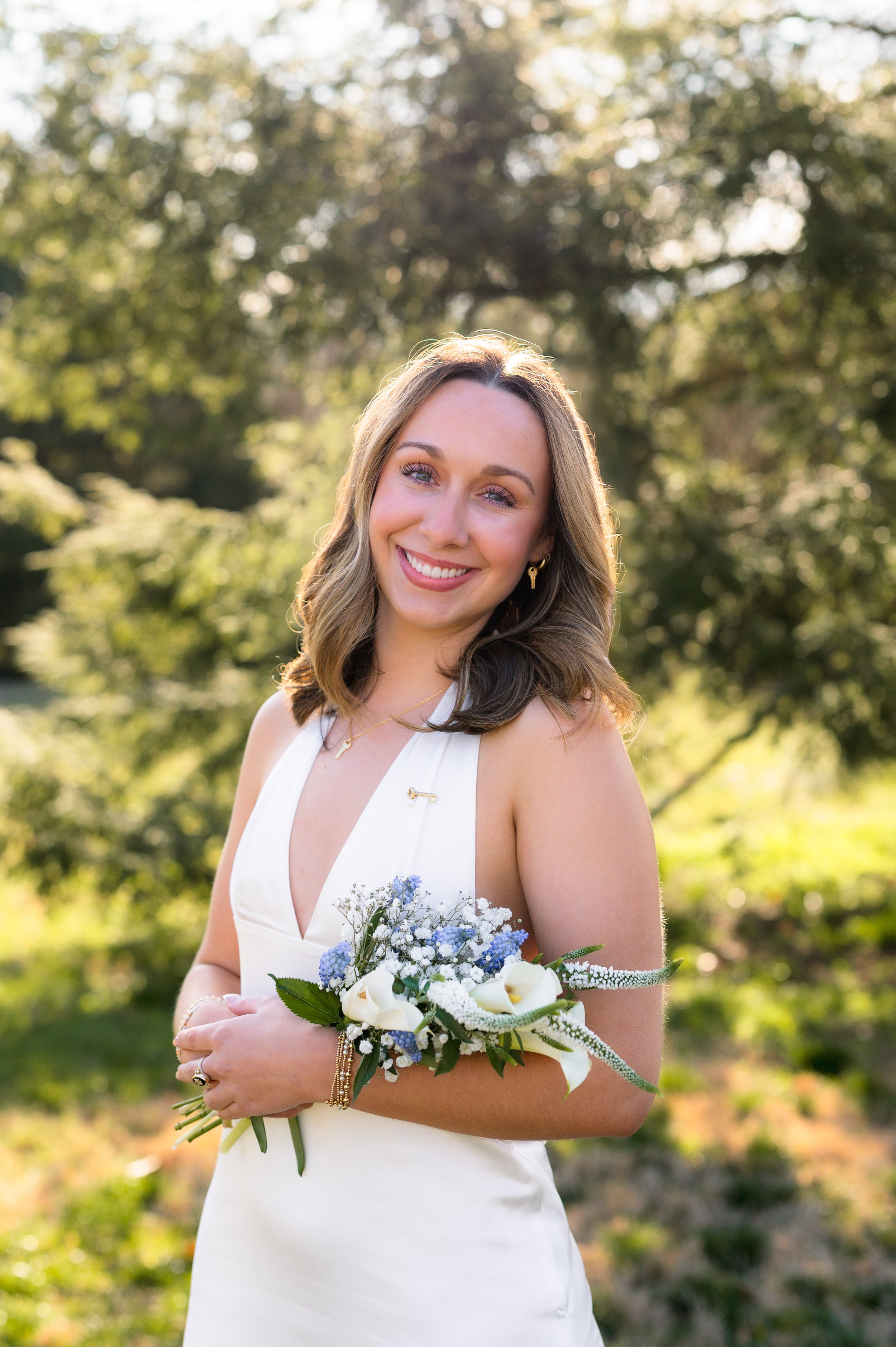 Young woman in a white dress holding a bouquet of white and blue flowers, standing outdoors in a sunny, green park with trees in the background.