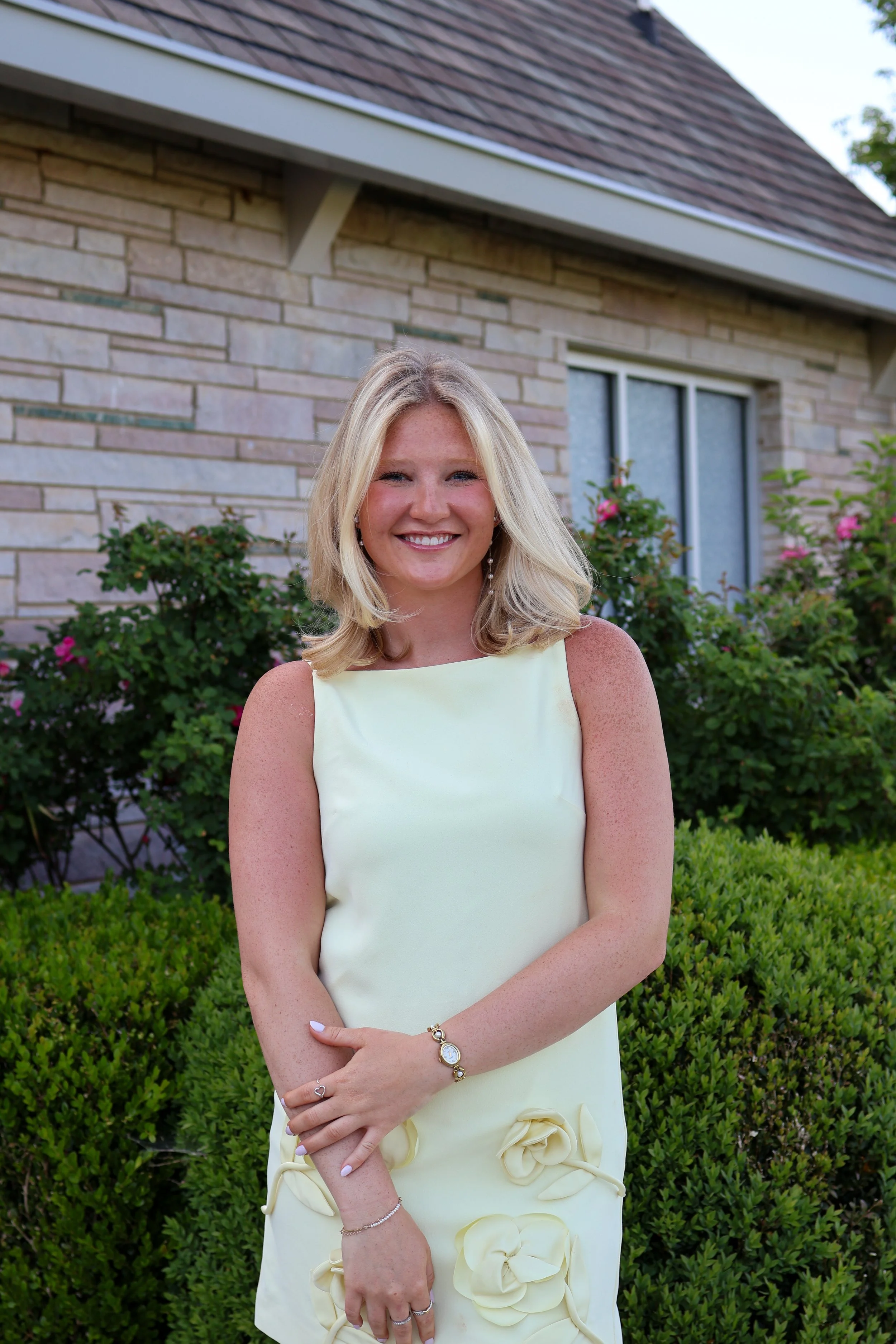 A woman with blonde hair smiling and standing outdoors in front of a house with a brick wall, behind bushes with pink flowers.
