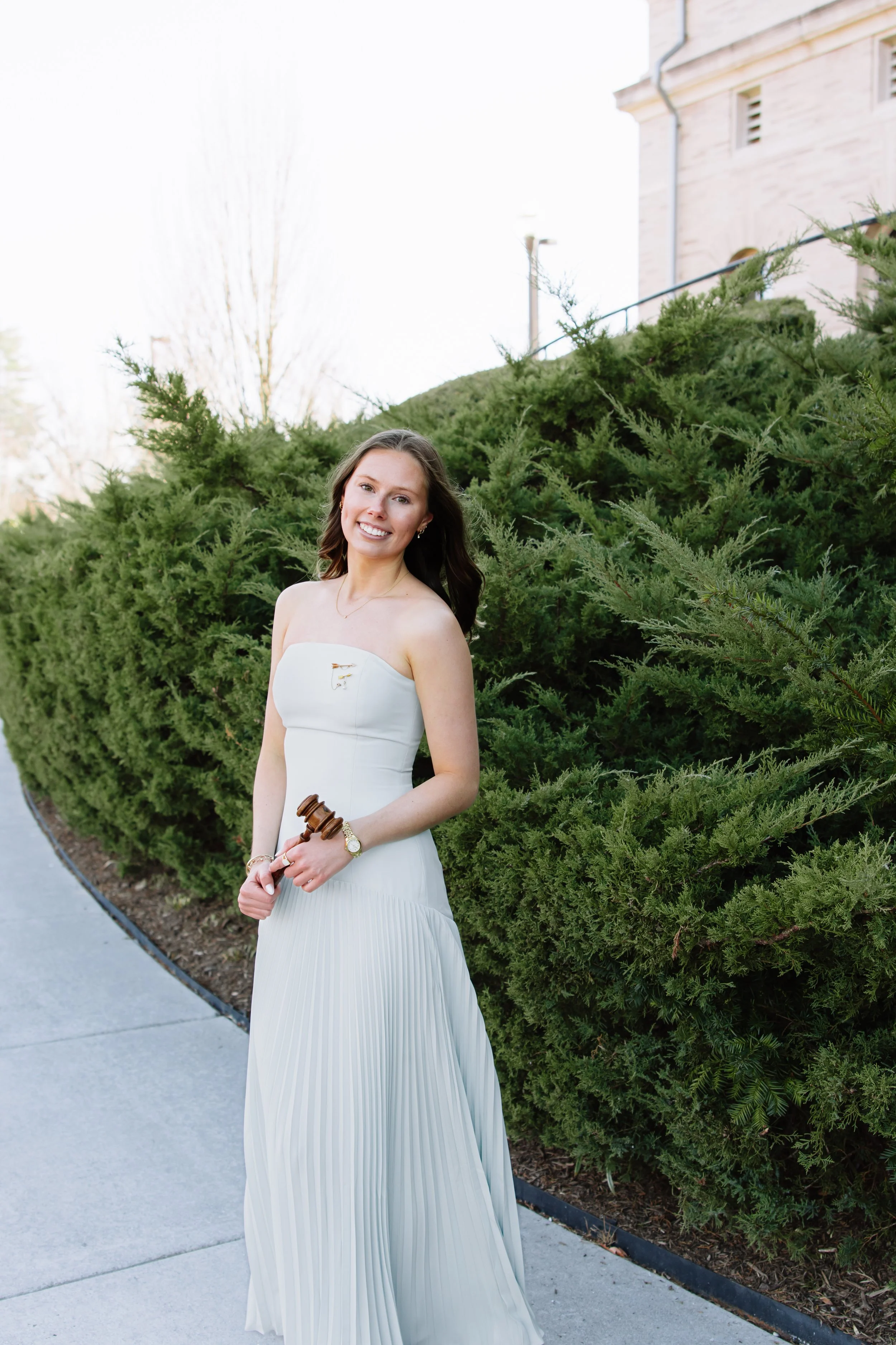 A smiling woman in a white strapless dress standing outdoors next to green bushes with a building in the background.