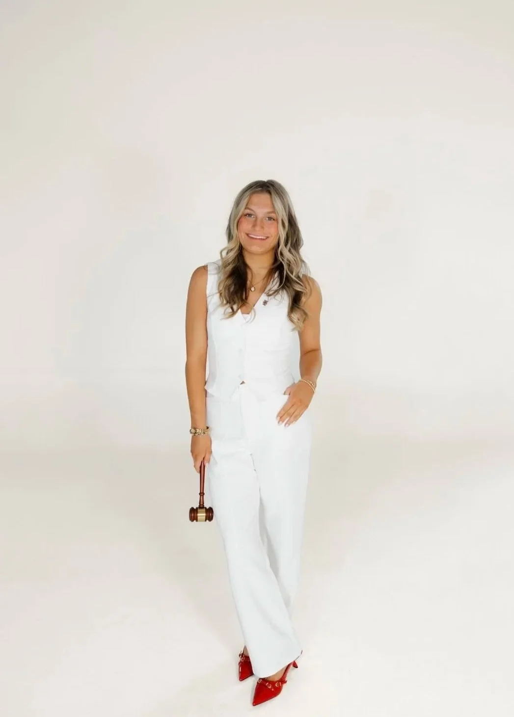 A woman with long wavy blonde hair smiling, dressed in a white sleeveless outfit, holding a small gavel, standing against a white background.