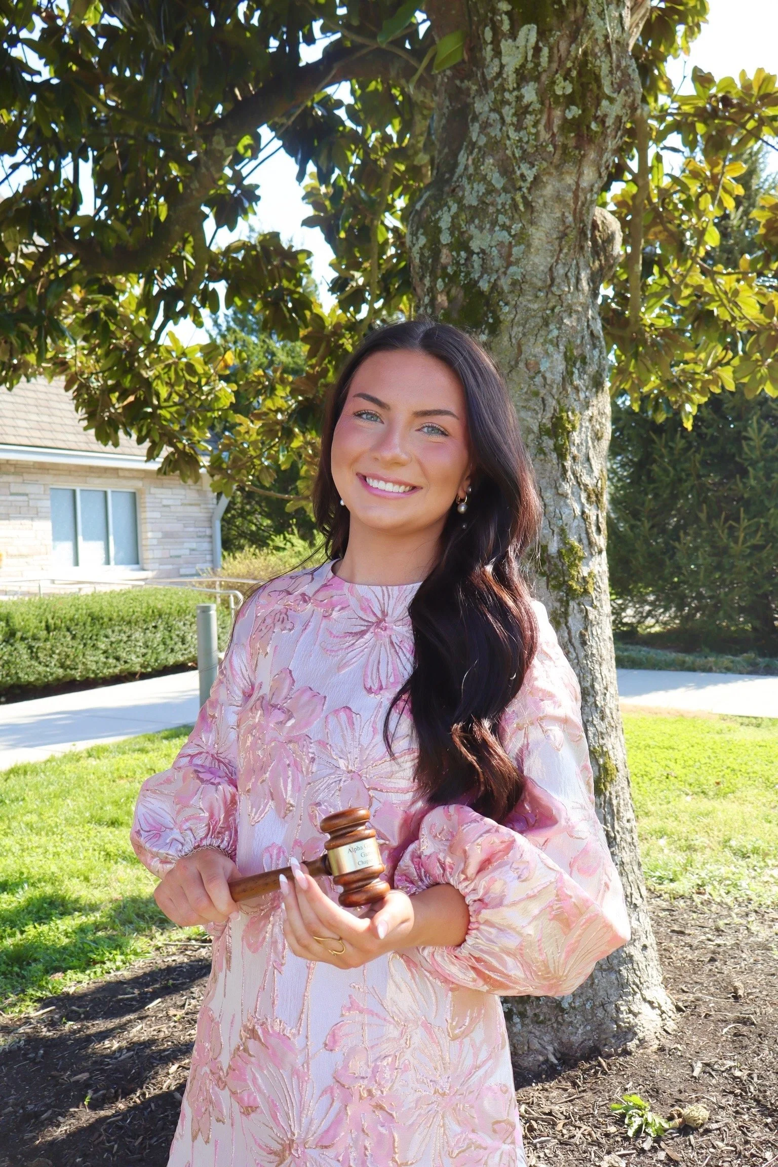 A young woman with long dark hair and a floral dress is standing outdoors next to a tree, smiling, holding a judge's gavel, in a sunny yard with a house in the background.