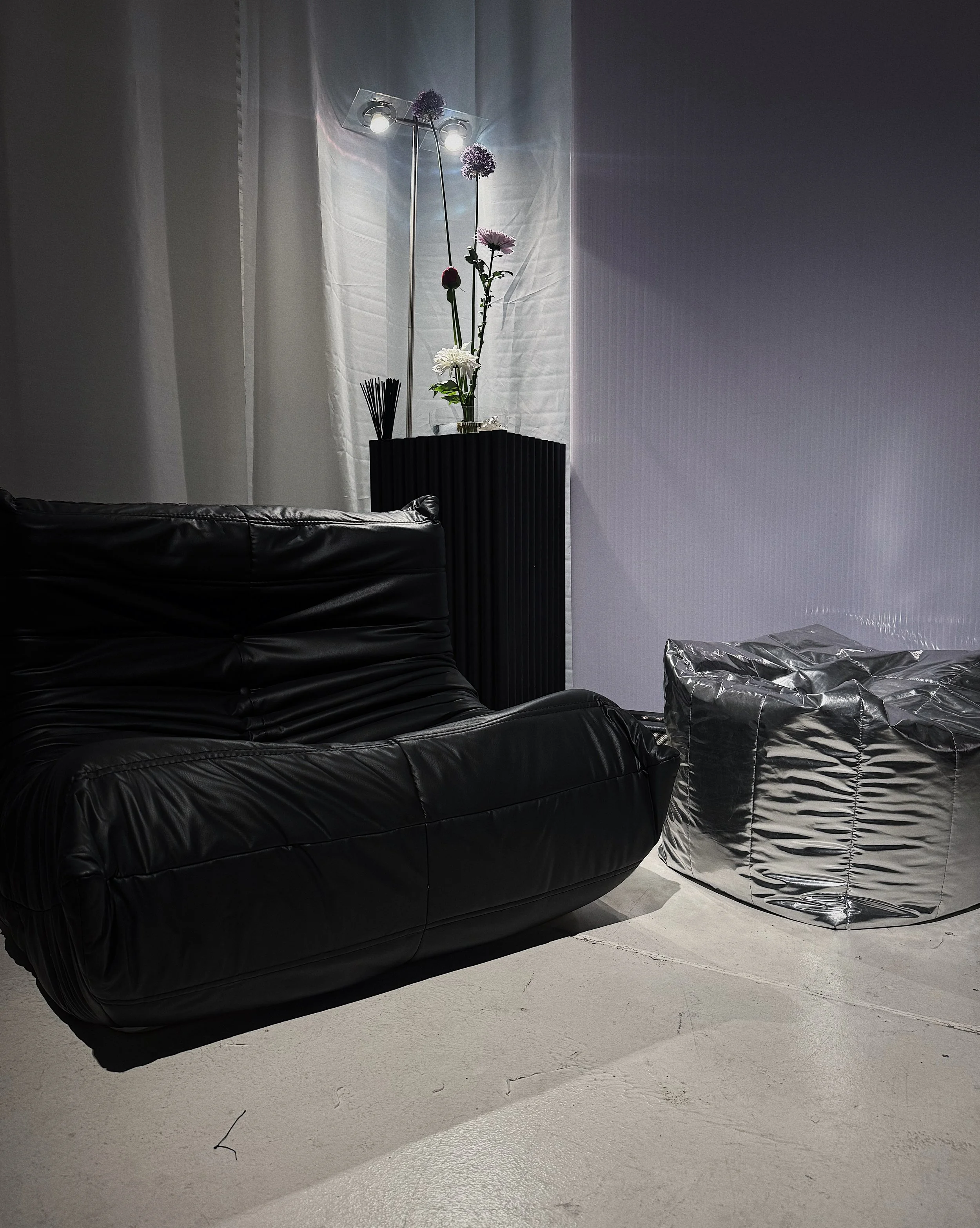 A black leather sofa, a metallic silver bean bag chair, and a black radiator with a vase of flowers on top in a room with plain white walls and a concrete floor.