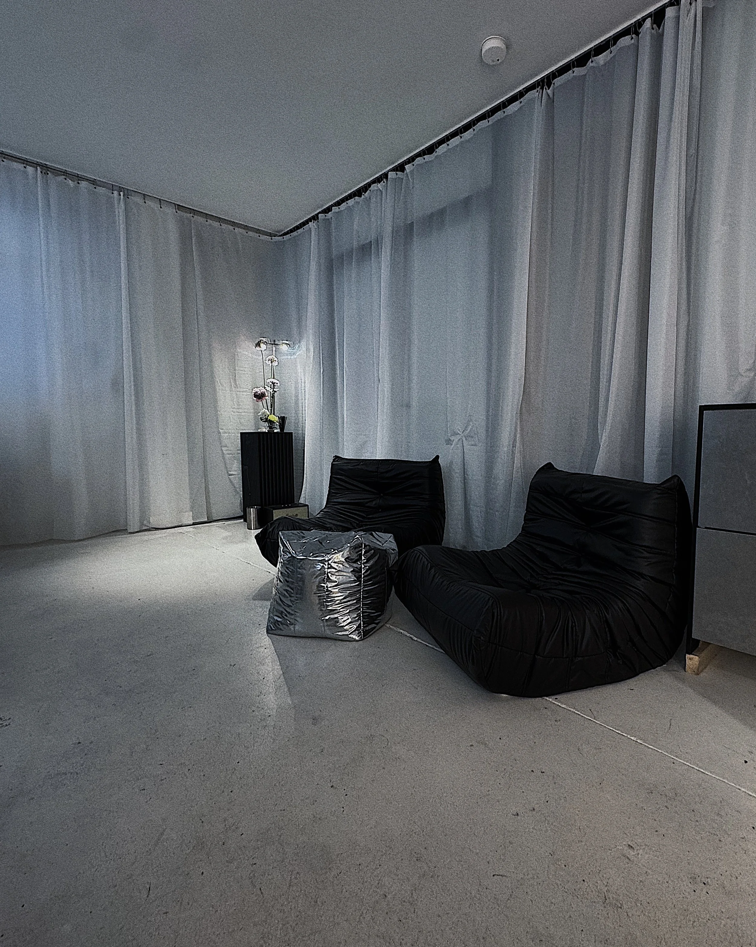 Interior of a room with gray curtains on the walls, a tall black flower vase with pink and white flowers, and two black leather chairs, one of which has a silver bag on it.