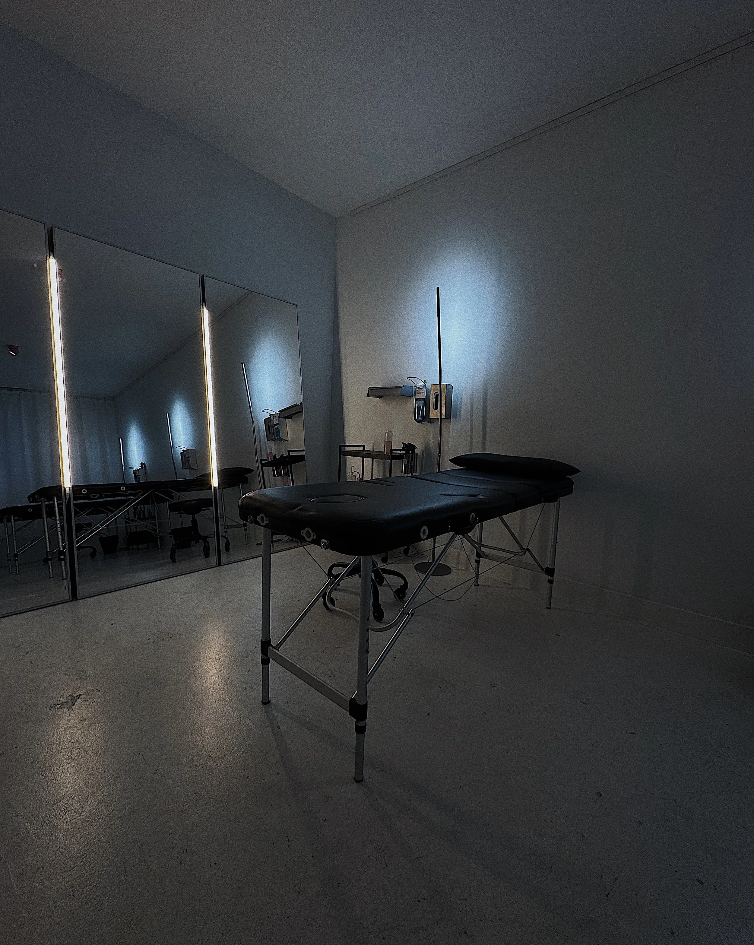 A dark-colored massage or acupuncture table in a dimly lit room with mirrored wall panels, small hanging lamps, and some shelves with equipment or supplies.