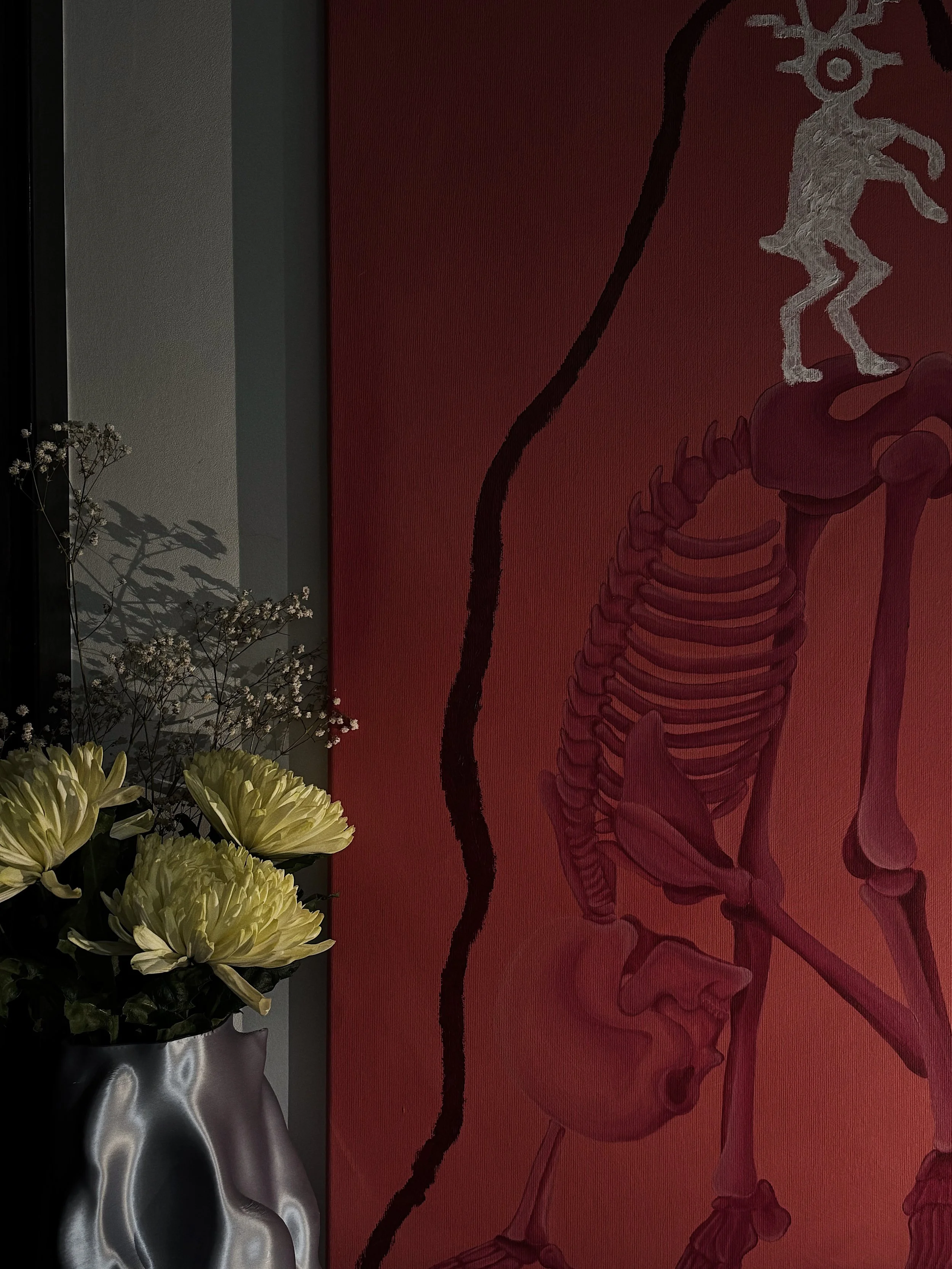 A framed flower arrangement with white flowers in a metallic vase on a table next to a wall art depicting a human skeletal and muscular system.